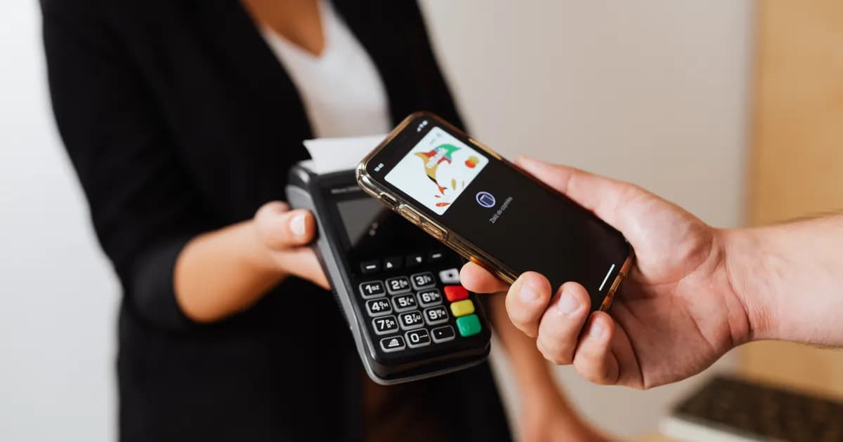 Mão segura smartphone sobre terminal de pagamento.