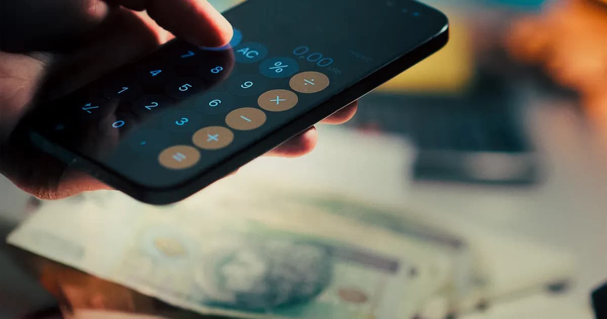 Pessoa consulta valores no celular ao lado de dinheiro.